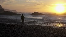 Silhouette of young man walk on rocky beach looking at beautiful wild sunrise in New Zealand ocean coast nature
