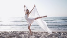Young beautiful ballerina dancing ballet on seashore with huge silk fabric fluttering in wind. Concept of tenderness, lightness, art and talent in nature