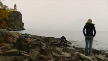a woman in a coat standing on rocky shore looking out at the ocean 
