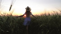 Freedom concept. Pretty child in the hat is running across the wheat field. Happy young girl running in the field at sunset. Happy kid playing in the wheat field on a warm summer day at sunset.