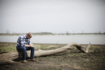 A young man praying while sitting on a log next to a lake bed.