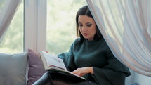 Beautiful charming woman in green clothing relaxing at home on cozy window sill and reading book. Lady smiling. Slow motion