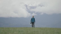 Young man walking up green hill in the mountains with clouds and mountains in background holding a camera 