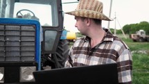 Farmer with laptop stood by tractor. Young farmer engineer with laptop standing on sun. This is a close up of a farmer using a computer on his tractor.