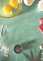 whisk, flour, measuring spoons, cracked eggs, bowl, and towel on a kitchen countertop 