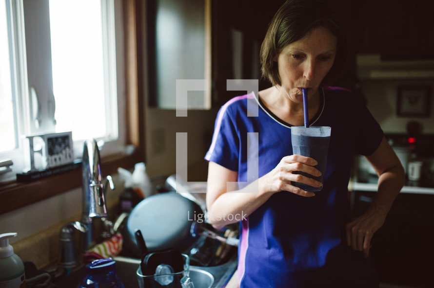 a woman drinking a smoothie in front of a sink of dirty dishes 