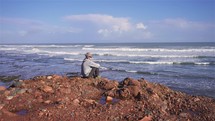 Man in hat and sunglasses sit and meditate on red rocky beach of ocean coast a in sunny summer morning Travel background
