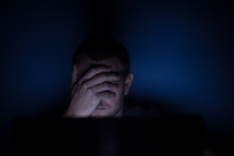 a man covering his eyes looking at a computer screen at night 