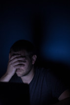 a man covering his eyes looking at a computer screen at night