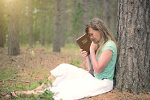 a woman sitting under a tree in a forest holding a Bible in prayer 