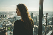 A woman looking from a window of a high rise building across a city.