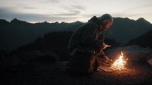 Man building and tending to fire on mountain at dusk (or sunset)