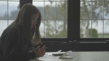 A young woman sits a table talking to someone across from her while she writes with a pen on paper.