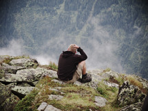 man sitting on the edge of a mountain scratching his head and enjoying the view