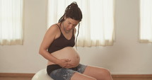Pregnant woman sitting on a yoga ball holding and rubbing her pregnant belly.