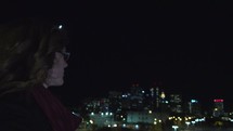 woman standing on a rooftop looking out at a parking lot at night