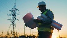 Architect Worker Checking Construction Project On Electric Tower. One Engineer Stands Near Power Lines At Sunset. Male Technician Is Operating A Documents Beside Electrical Transmission Lines.