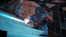 Unrecognizable man in protective clothing and mask at construction factory is engaged in welding work. Concept of building and work with metal. Sparks from modern machine