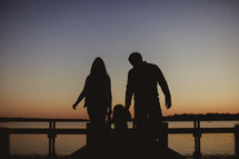 silhouette of a mother and father holding hands with their daughter walking on a dock