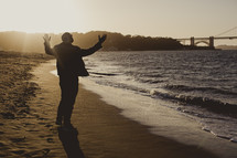 man standing on a beach in front of the ocean with his hands raised in praise