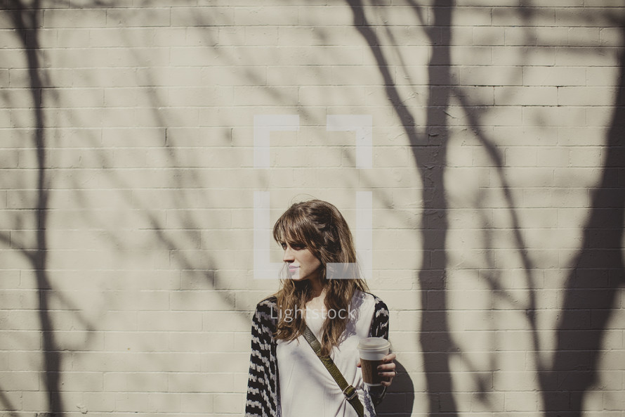 woman standing in front of a brick wall holding a coffee mug and the shadows of tree branches