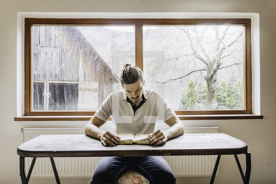 a man sitting at a desk reading a Bible with a farm scene in the window behind him 