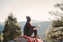 a woman sitting on a blanket on a rock drinking coffee 