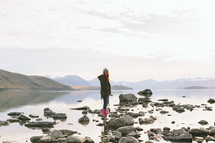 A girl stands on rocks at a lake's edge.