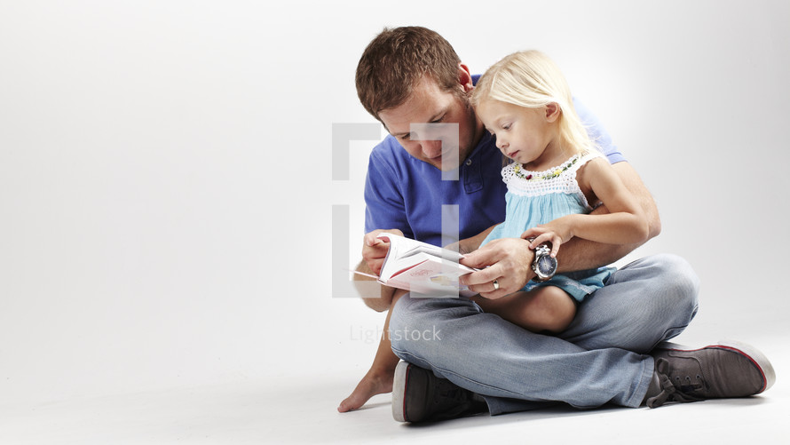 A dad reads a Bible with his young daughter in his lap.