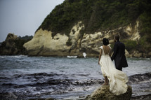a bride and groom standing on a rock along a shore 