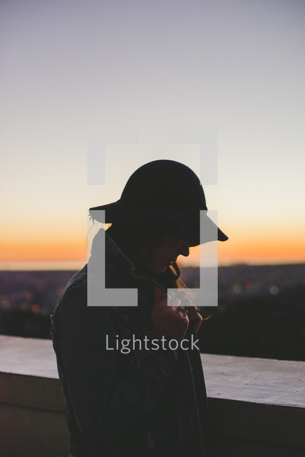 silhouette of a woman in a hat standing outdoors at dusk 