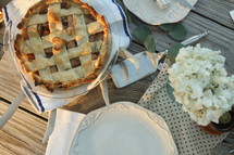 Bouquet of flowers, plates, and an apple pie on a picnic table outside.