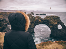 a woman in a coat looking out at a rock arch formation in the ocean 
