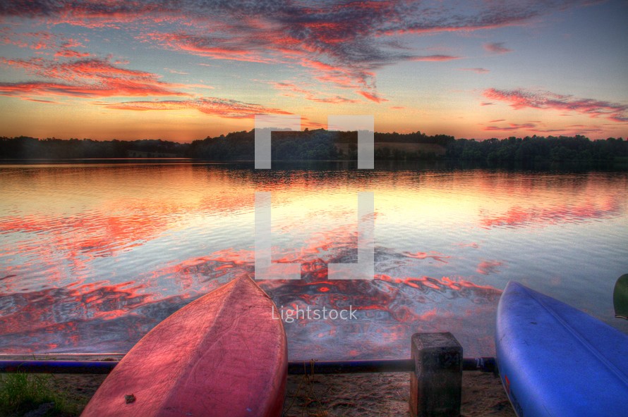 pink and purple clouds reflecting on lake water in front of two flipped over  boats resting on a shore