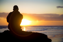 man sitting at a rock at sunset looking out at the ocean 
