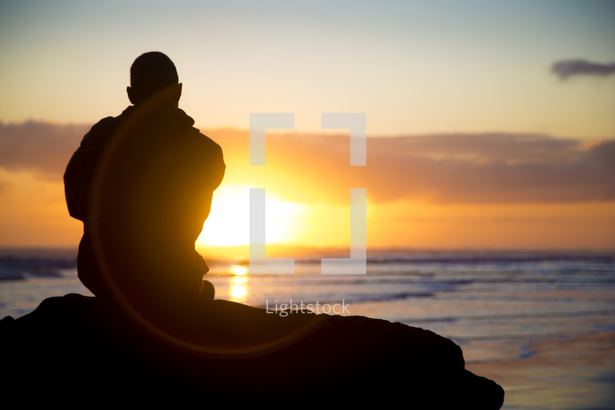 man sitting at a rock at sunset looking out at the ocean 