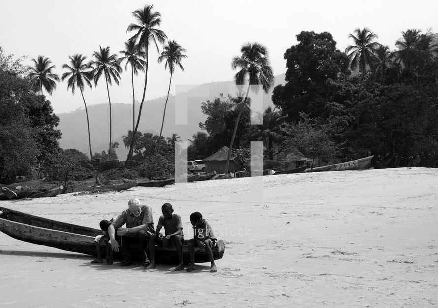 A man and children sit on a canoe on a beach in a tropical setting.