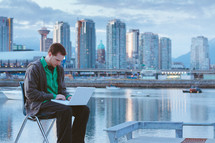 Man sitting on a chair with a laptop computer by a bay with a boat, near a city skyline.