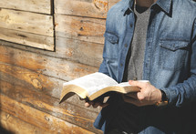 a young man reading a Bible in front of a wall of wood boards 