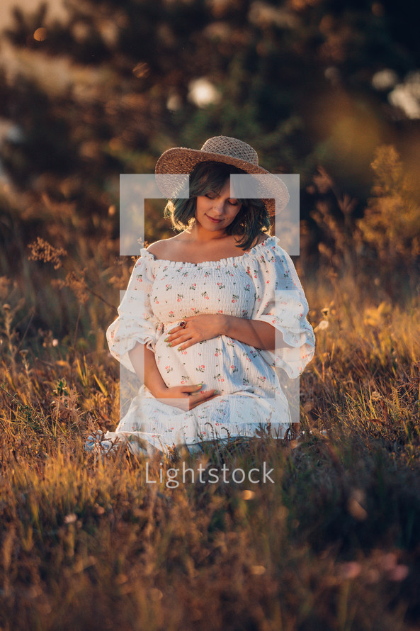 Happy Mother Carrying Child, Expecting For Baby. Amazing Motherhood. Pregnant Woman in beautiful dress and straw hat sitting on grass at nature rural meadow. High quality 