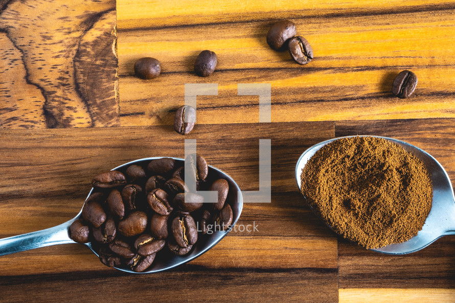 Spoons with coffee powder and coffee beans, laying down on a wooden background.