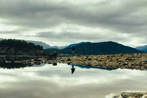 reflection in lake water of a man walking along a shore 