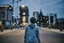 a teen boy standing in the middle of a road looking back at a city 
