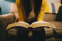 A woman sitting on a couch reading a Bible. 