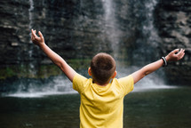 a child with outstretched arms standing in front of a waterfall 