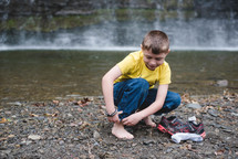 a child taking off his shoes to roll up his pants and play in a stream 