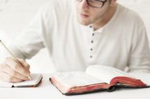 Man writing in a journal with a pencil next to an open Bible.