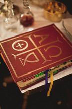 A religious book known as the Lectionary with the Alpha and Omega symbols on the front cover - also contains tabs and multiple bookmarks.