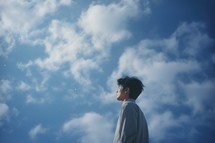 Faith. Heavenly background. Young asian man on blue sky background with white clouds and copy space