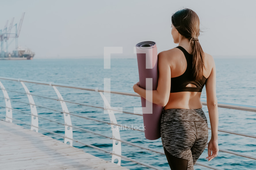 Young sports woman walking to do morning stretching practice. Girl holding yoga mat. Sea or ocean background. Summer days. 
